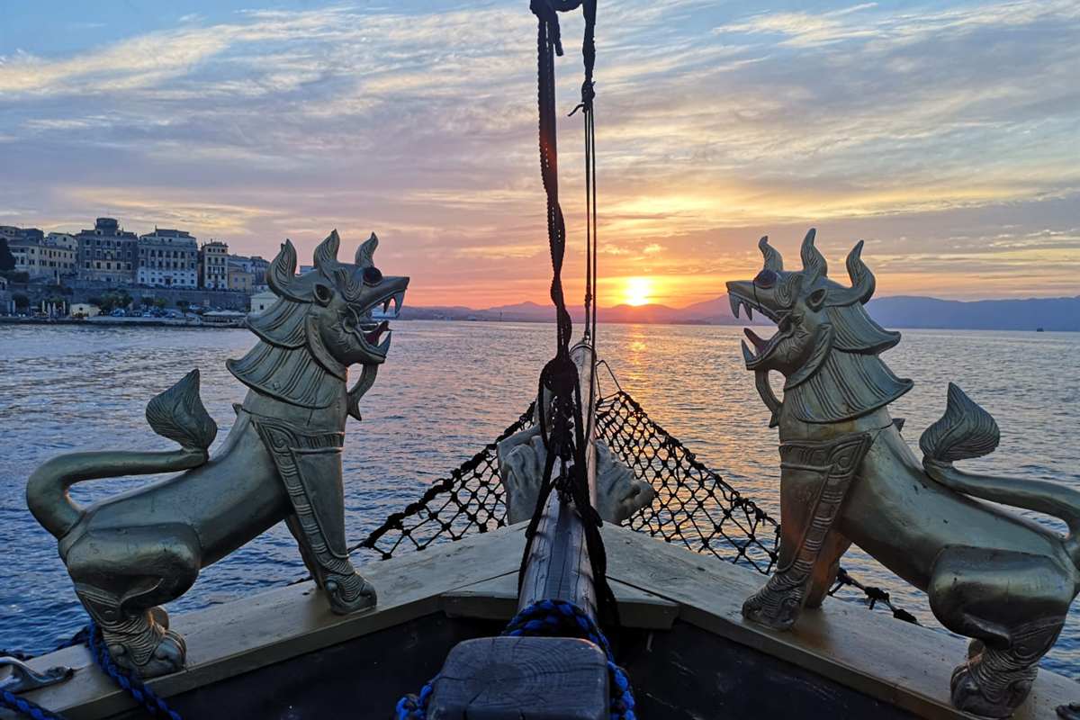Corfu pirate ship: two golden lion statues on the bow of a wooden pirate-style ship with a black rope net between them, facing a low orange sun over calm sea, with the pastel buildings of Corfu Town along the shore and layered blue hills beyond