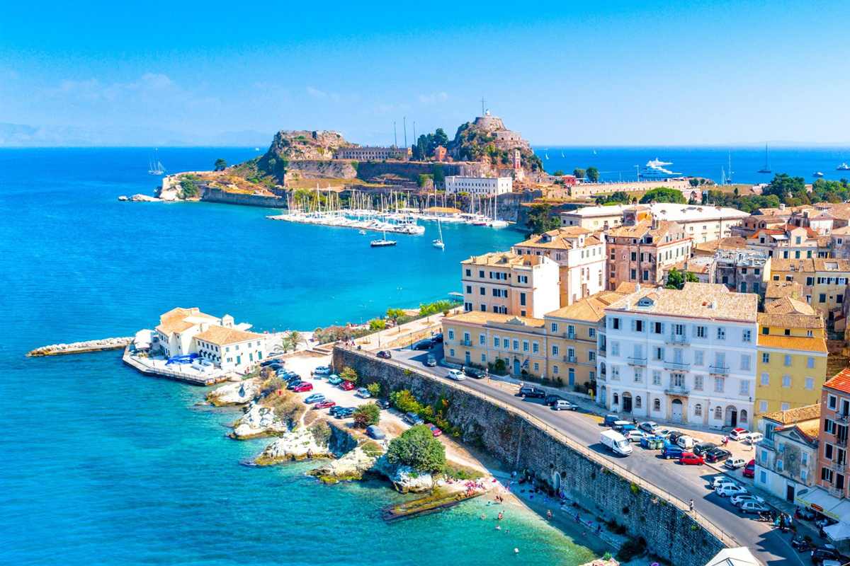 Corfu shore excursion: the Old Fortress of Corfu on its rocky headland above a marina of white sailboats, beside the pastel orange, cream and white houses of Corfu Old Town and a sea wall road, with clear turquoise water under a bright blue sky