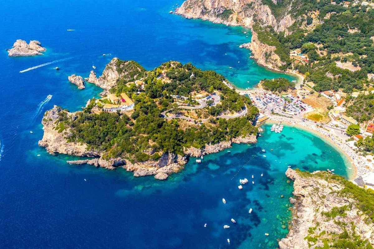 Corfu shore excursion: aerial view of the green headland of Paleokastritsa with white buildings and yellow roofs on top, a curved sandy beach on the right and small white boats on turquoise and deep blue water below pale rocky cliffs
