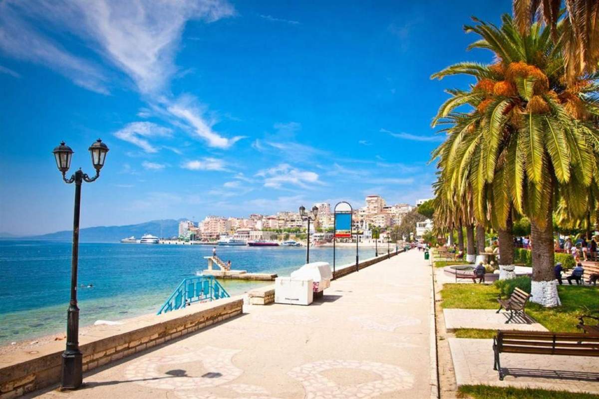Corfu to Albania day trip: the sunny seafront promenade of Saranda with black street lamps, tall palm trees and wooden benches, calm blue water on the left and pale apartment blocks and moored boats along the harbour under a bright blue sky