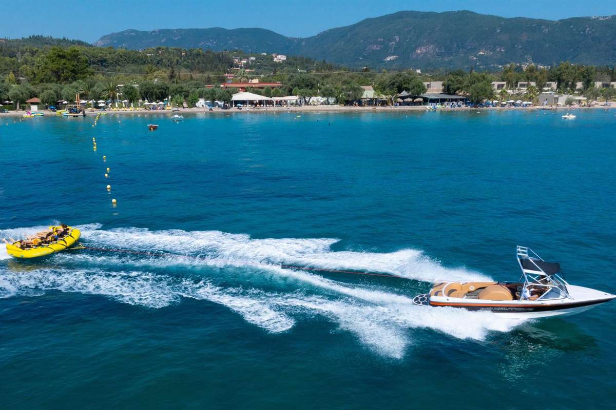 Corfu watersports: a white speedboat towing a yellow inflatable ride with riders lying on it, leaving a wide white wake across deep blue water off a sandy beach with a white hotel, palm trees and green hills