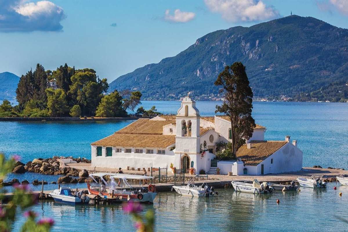 Corfu watersports: the white Vlacherna monastery with a bell tower and orange tiled roofs on a small pier beside Pontikonisi, a tree-covered islet, in calm blue water with small boats and a green mountain behind under a blue sky