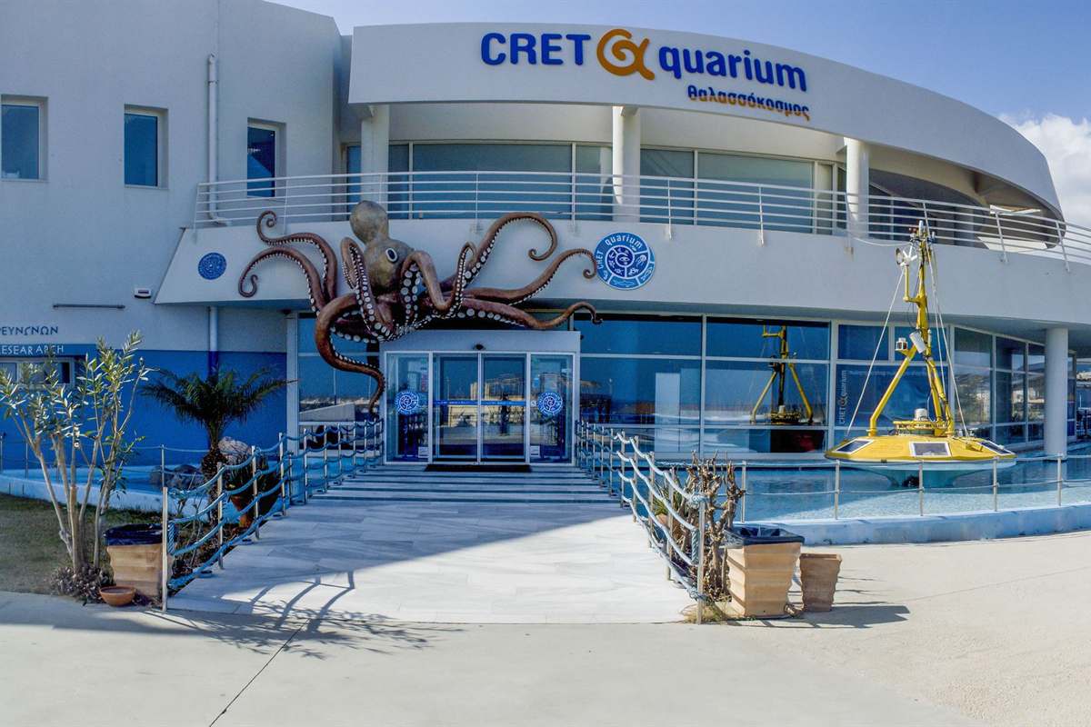 Cretaquarium tickets: the white curved entrance building of the aquarium at Gournes with a blue sign on its roof edge, a large brown octopus sculpture with white suckers above the glass doors, marble steps with rope railings and a yellow research buoy beside a shallow pool under a blue sky
