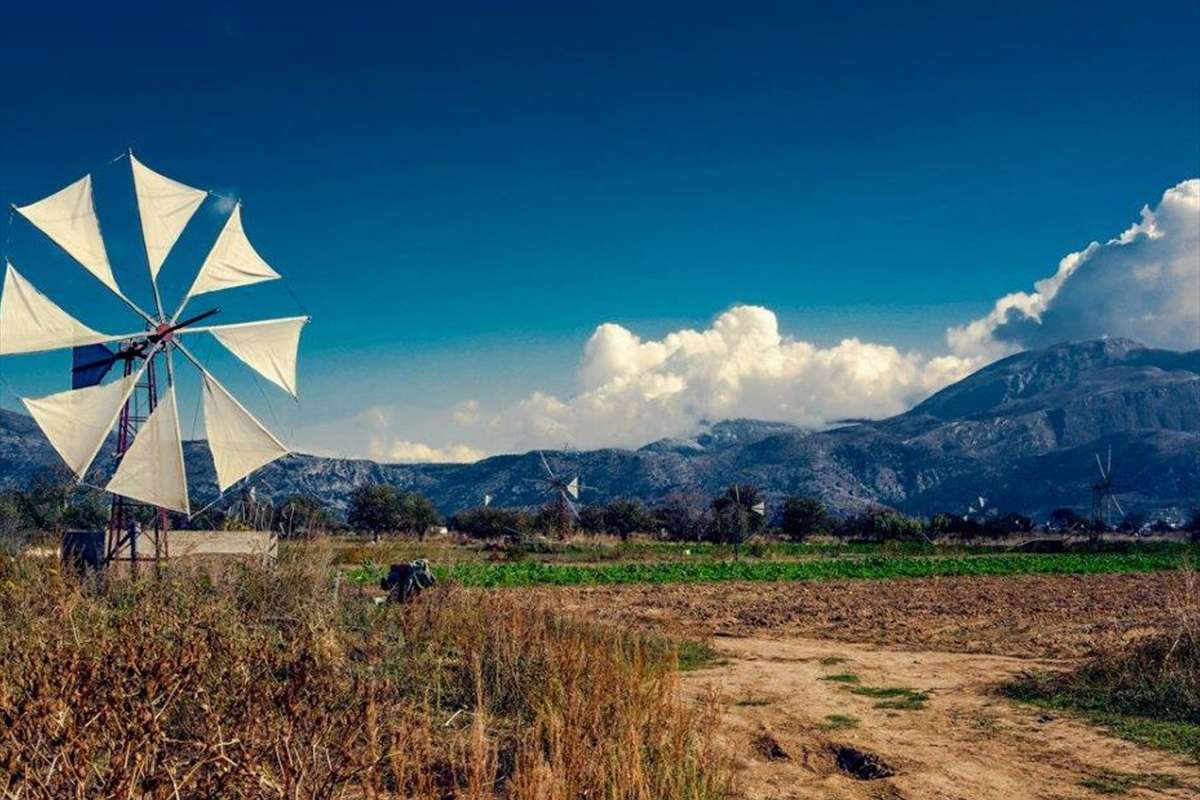 Crete jeep safari: a white-sailed windmill with ten cloth sails standing in a farmed plain on the Lasithi Plateau, with more windmills far behind it, dry grass and a dirt track in front, rugged mountains beyond and a deep blue sky with large white clouds