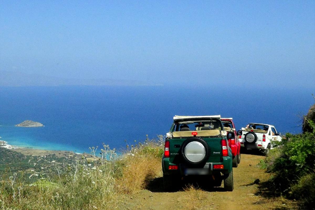 Crete jeep safari: three open jeeps, one green, one red and one white, driving in a line along a narrow dirt track on a mountain slope, with wild grass and flowers in front, a deep blue Cretan bay with a small rocky islet far below and a hazy blue sky above