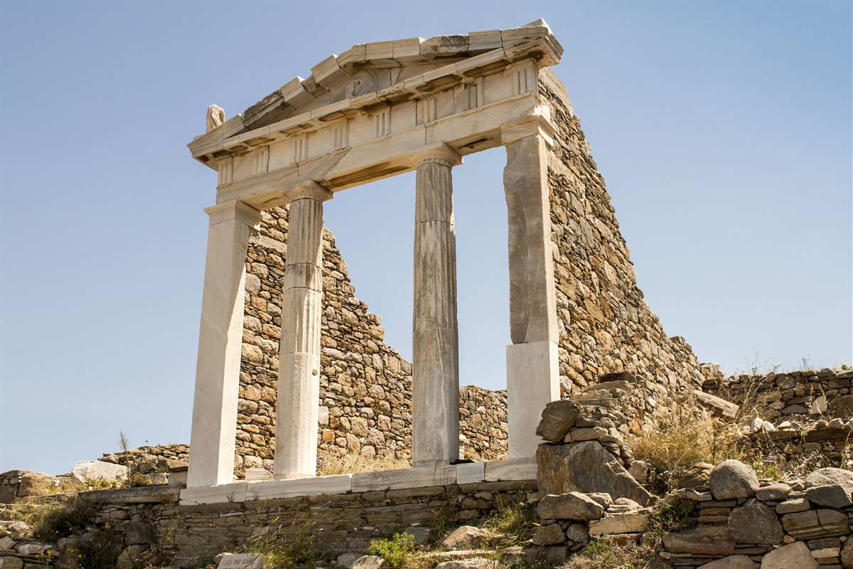 Delos tour from Mykonos: the restored white marble front of the Temple of Isis on Delos, three fluted columns holding a triangular pediment against a rough stone wall and tumbled stones, under a clear blue sky