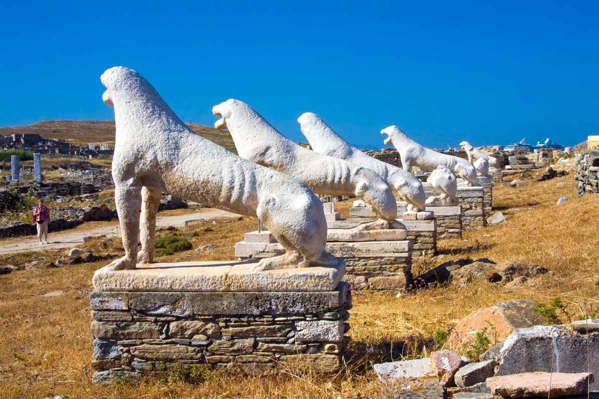 Delos tour from Mykonos: a row of weathered white marble lion statues with open mouths on stone pedestals on the Terrace of the Lions, with dry golden grass, low ancient ruins, a visitor on the path at left and a deep blue sky behind