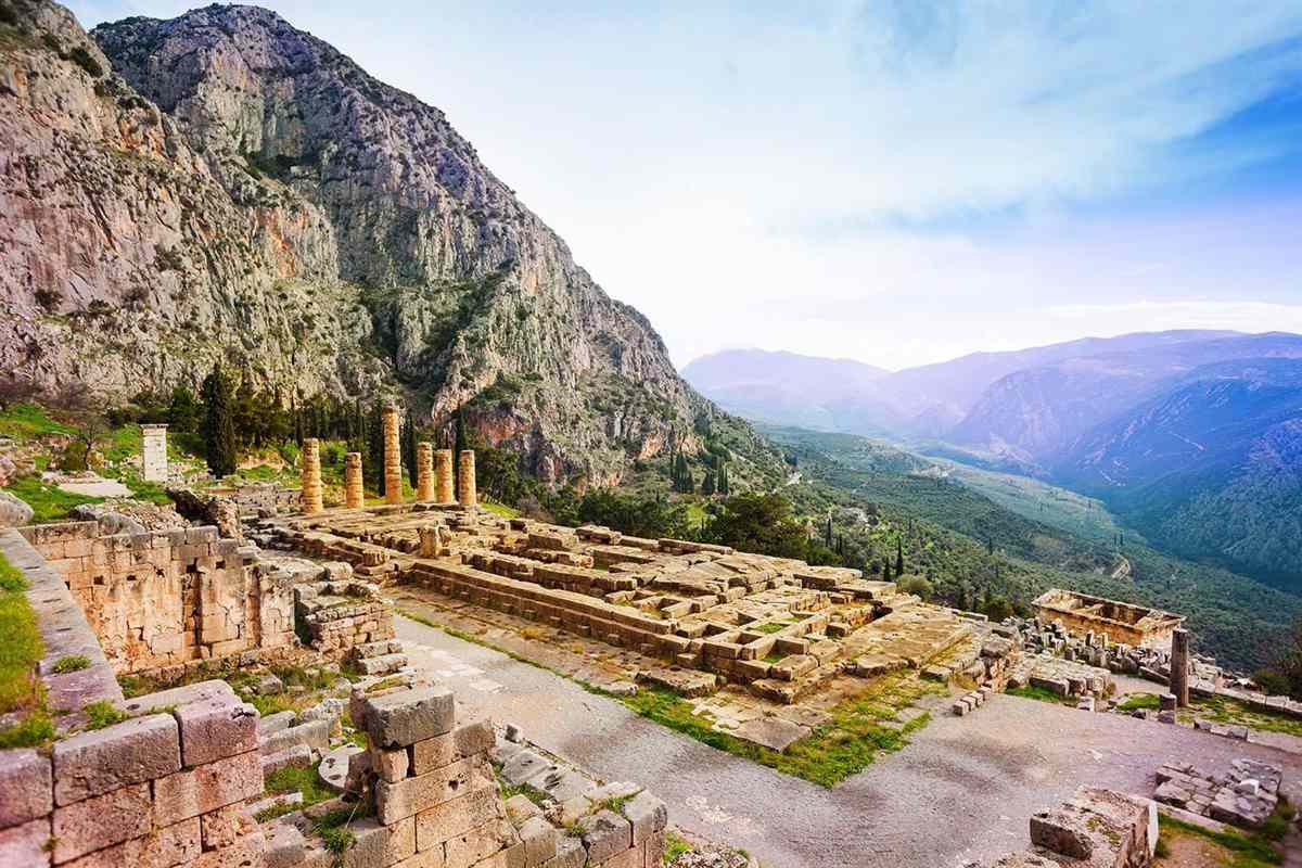 Delphi day trip from Athens: six standing golden columns of the Temple of Apollo above its ruined stone terrace at Delphi, with a steep limestone cliff of Mount Parnassus on the left and a green olive valley fading into hazy blue mountains on the right