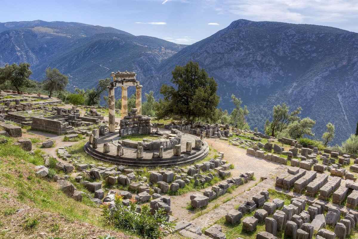 Delphi day trip from Athens: the circular Tholos of Athena Pronaia with three restored Doric columns on its round marble platform, surrounded by fallen stone blocks, a dark cypress tree and olive trees, with a wide mountain valley behind under a blue sky