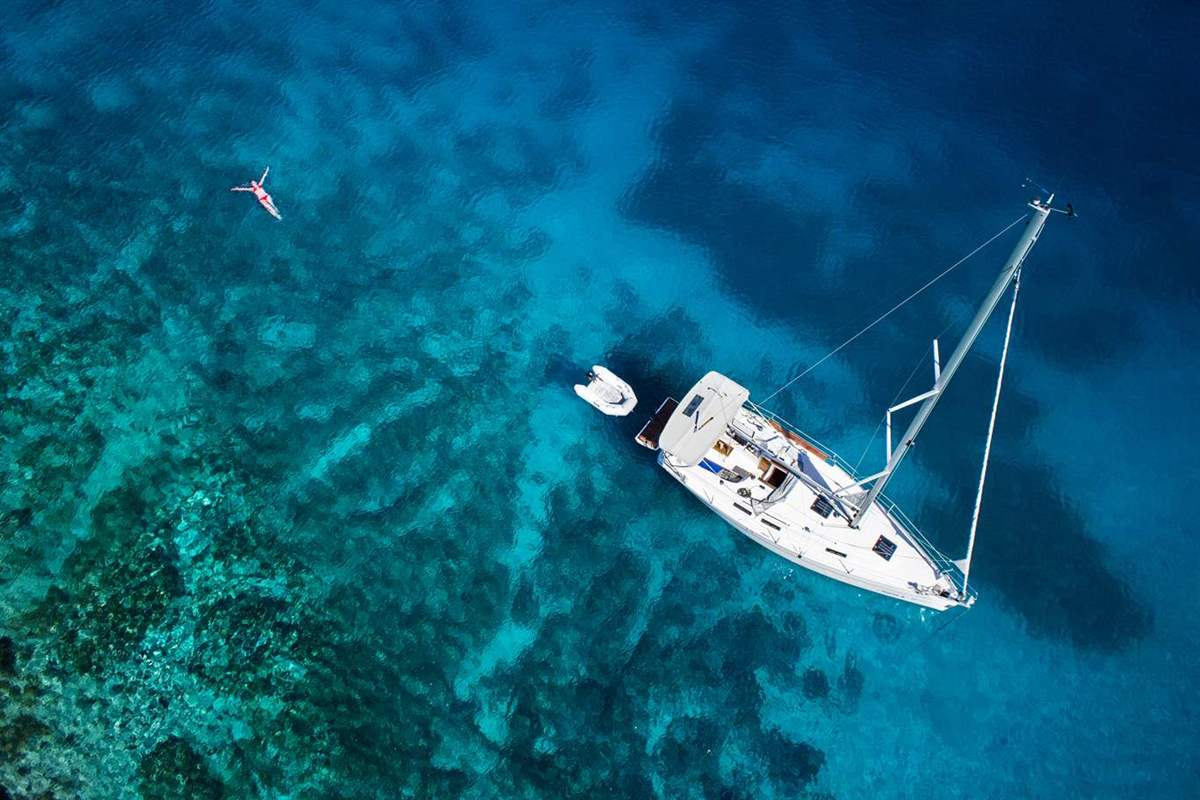 Dia island cruise from Heraklion: an aerial view of a white sailing yacht and its small dinghy at anchor over deep blue and turquoise water with a rocky seabed, and a lone swimmer floating on the left