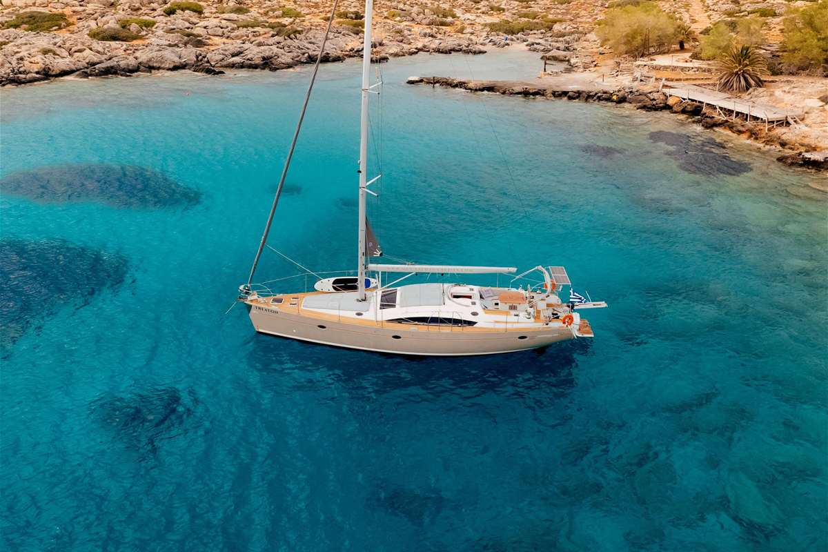 Dia island cruise from Heraklion: a cream-hulled sailing yacht anchored in a clear turquoise cove, seen from above, with a rocky shore, low green scrub, a palm tree and a small wooden jetty beyond