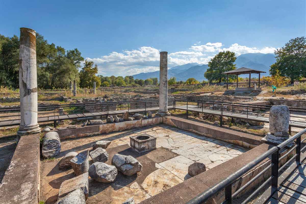 Dion and Mount Olympus day trip: two tall white marble columns and a cracked stone-paved sanctuary floor with fallen blocks and a square well at the ancient site of Dion, wooden walkways and a small pavilion beyond, and blue ridges of Mount Olympus under a clear blue sky