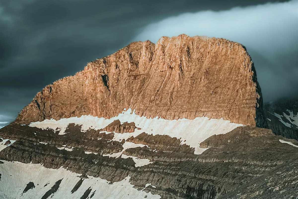 Dion and Mount Olympus day trip: a sunlit orange limestone summit wall of Mount Olympus above bands of grey rock and white snow patches, under dark storm clouds