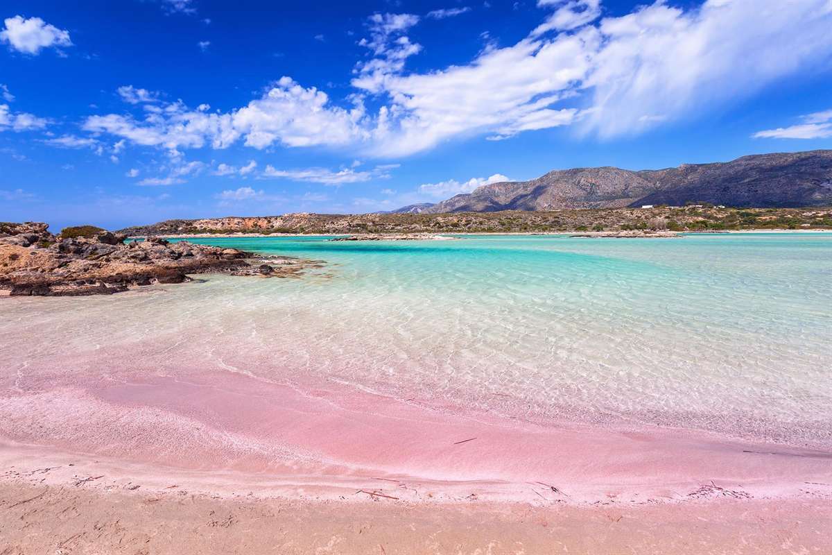 Elafonisi day trip from Chania: a stretch of pink sand at the edge of the Elafonisi lagoon, with clear turquoise shallows, a dark rocky outcrop at left and a low mountain ridge under a deep blue sky with white clouds