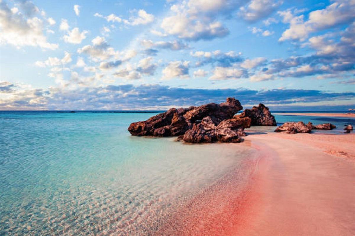 Elafonisi day trip from Chania: a band of pink sand curving along the shore beside clear turquoise water, with a cluster of dark brown rocks standing in the shallows under a sky of scattered white clouds