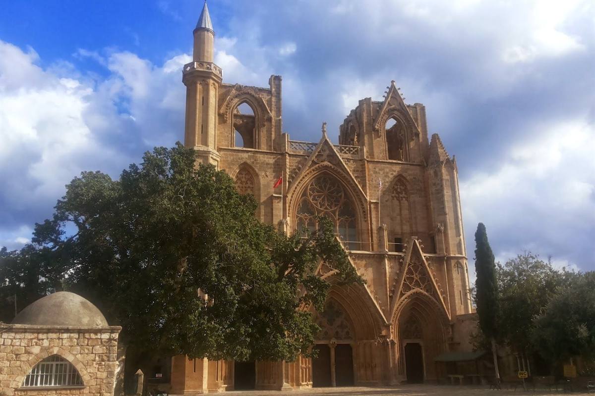 Famagusta ghost town tour from Ayia Napa: the Gothic stone front of the Lala Mustafa Pasha Mosque in Famagusta, with a tall minaret on the left, a large rose window, pointed arches and a big green tree and small domed building in front, under a blue sky with dark clouds
