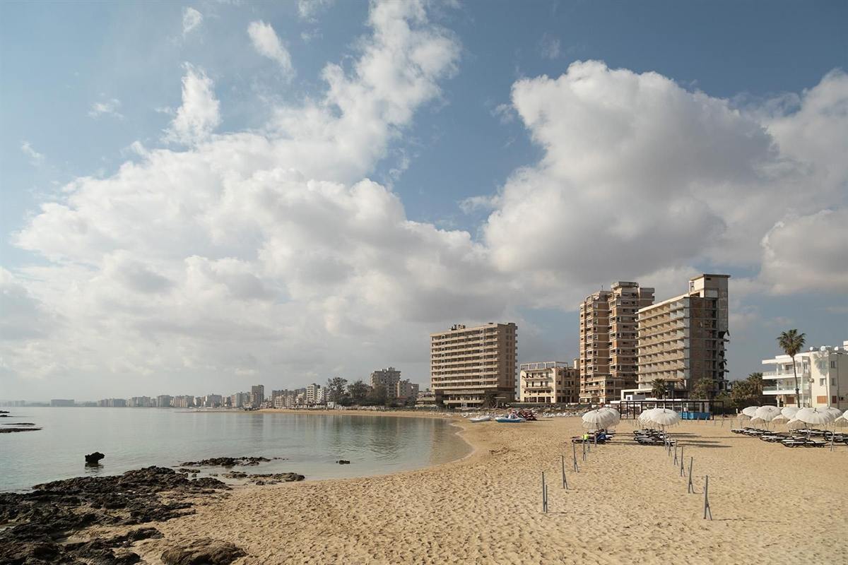 Famagusta ghost town tour from Ayia Napa: a curve of golden sand and calm sea in front of the empty high-rise hotel blocks of Varosha, with a long line of apartment buildings along the distant shore, rocks in the foreground and large white clouds in a blue sky