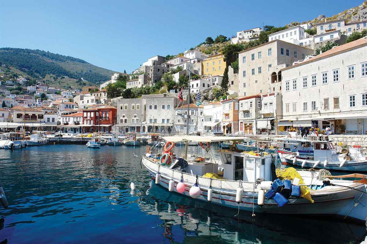 Hydra day trip from Athens: a white and blue wooden fishing boat with orange life rings and floats moored in the deep blue water of Hydra harbour, with grey stone mansions, red and yellow houses and green hills rising behind it under a clear blue sky