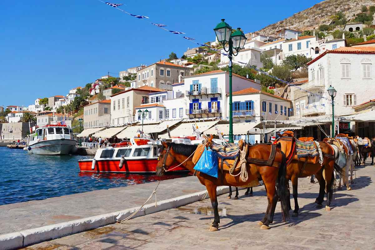 Hydra day trip from Athens: saddled horses standing on the paved stone quay of Hydra harbour beside a green lamp post, with a red and white boat on the water and white and stone houses with blue shutters climbing the hill behind, strung with blue and white flag bunting