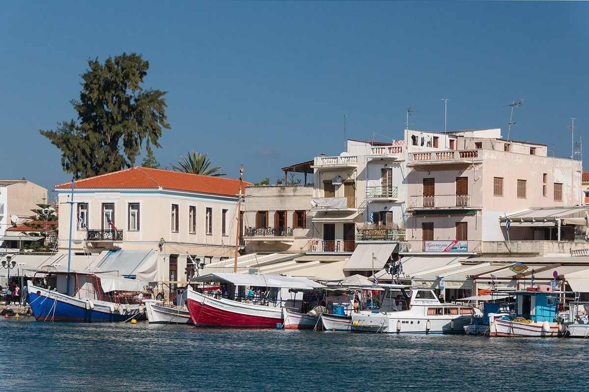 Hydra Poros Aegina day cruise: painted fishing boats moored along the Aegina harbour front below pastel houses and cafe awnings