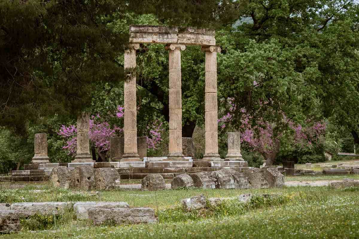 Katakolo to Olympia: the three slender fluted columns of the round Philippeion at Ancient Olympia carrying a stone beam, with pink blossom trees and thick green leaves behind them and fallen stone blocks lying in the grass in front