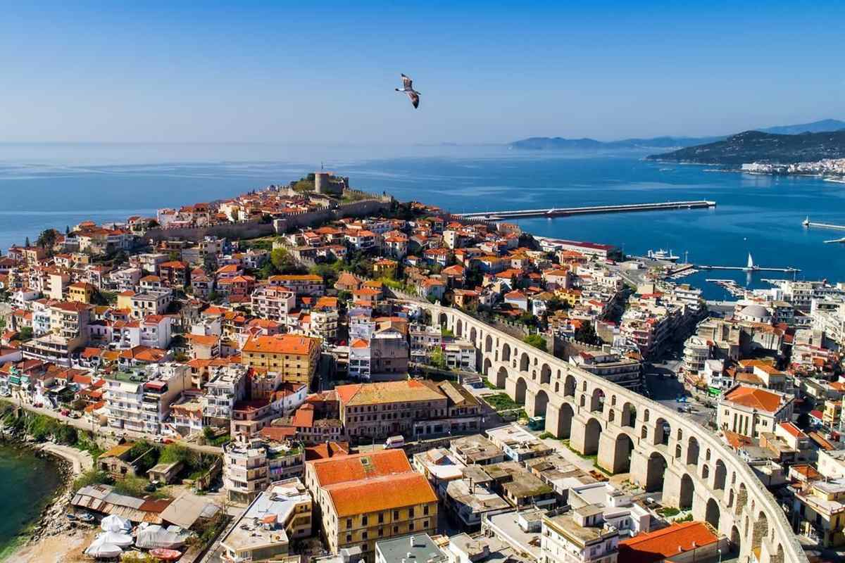 Kavala and Philippi day trip: aerial view of the old town of Kavala on its headland, with the stone castle at the top, the long arched Kamares aqueduct curving past terracotta roofs and a harbour pier on deep blue water under a clear sky