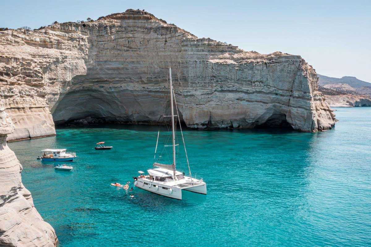 Kleftiko boat tour Milos: a white catamaran anchored in the turquoise Kleftiko bay under tall pale cliffs and sea caves, with swimmers and small boats nearby