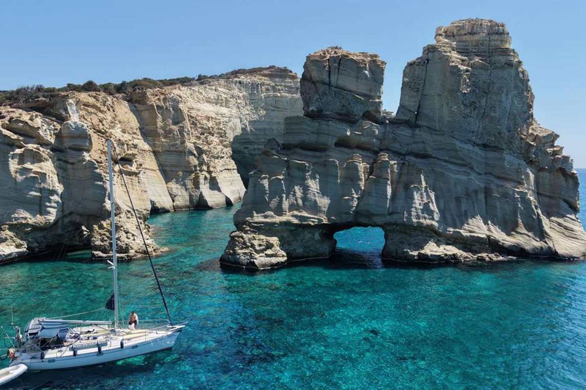 Kleftiko boat tour Milos: a sailboat anchored in clear turquoise water beside the white rock stacks and sea arch of Kleftiko on a sunny day