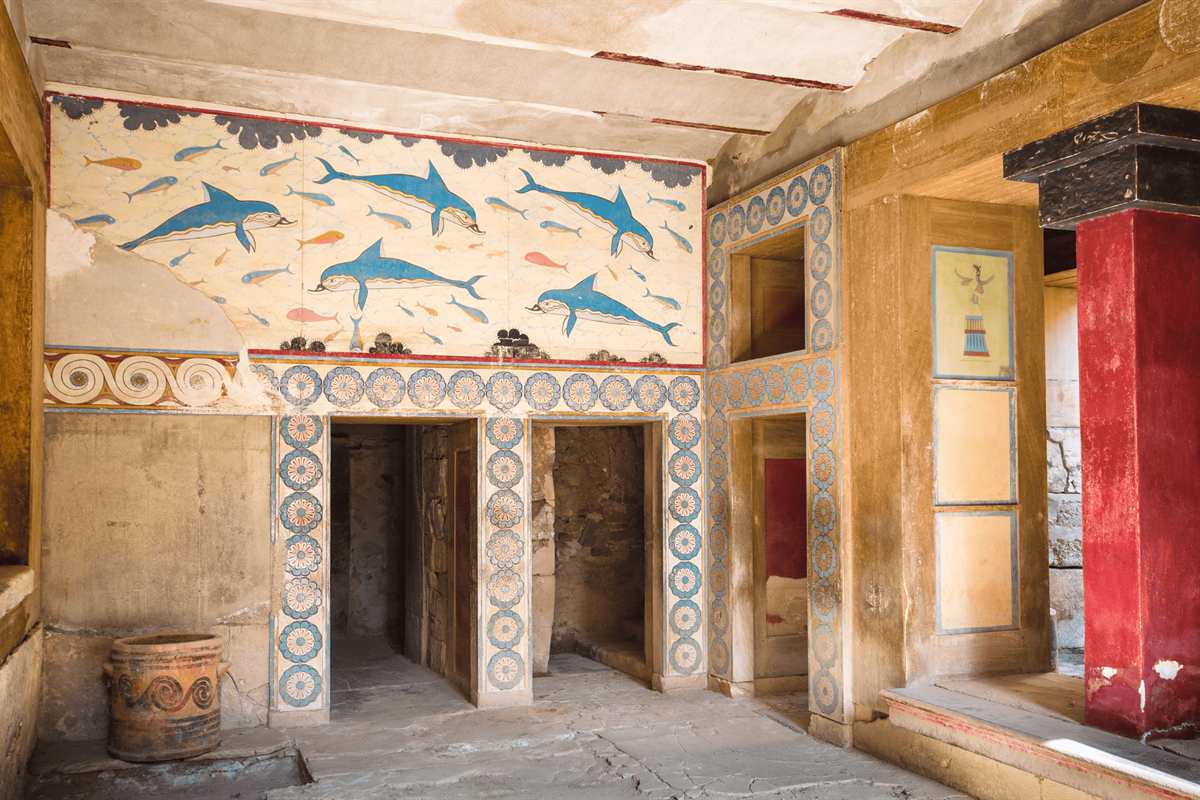 Knossos palace tickets: a painted room in the Palace of Knossos with a frieze of blue dolphins and small fish above two doorways framed by blue and orange rosette patterns, a red column on the right and a spiral-decorated clay jar on the floor