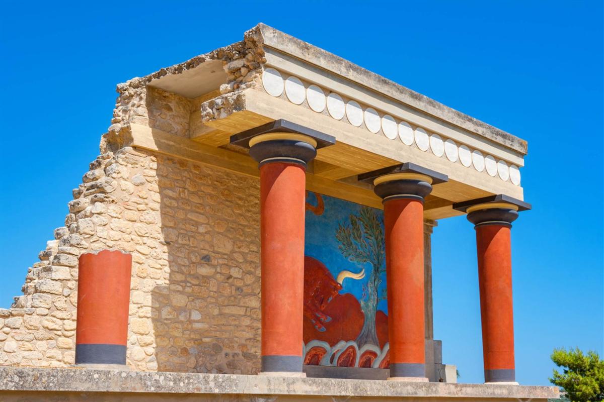 Knossos palace tickets: the restored north entrance of the Palace of Knossos with three tapering red columns, a painted bull fresco on the wall behind them and a pale stone roof edge with white discs under a clear blue sky
