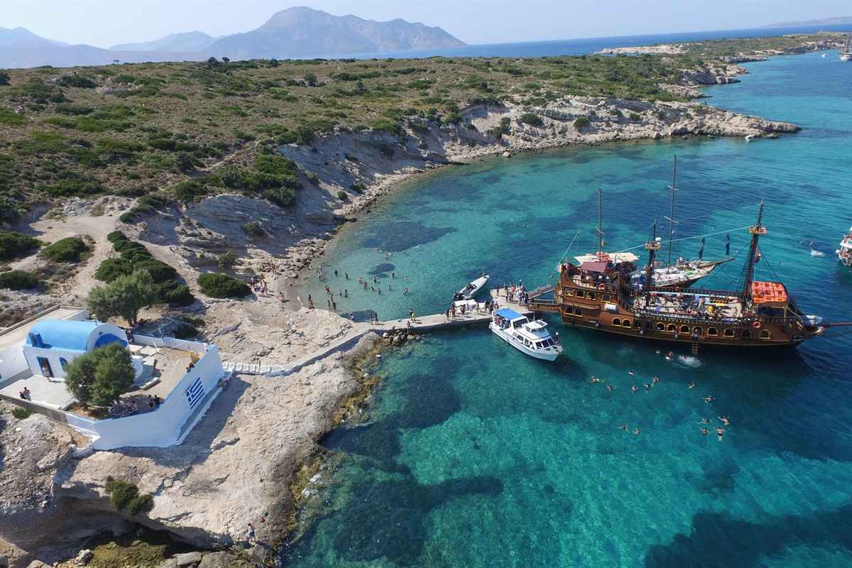 Kos 3 islands boat trip: aerial view of a brown wooden pirate-style cruise ship moored at a stone jetty in a turquoise bay beside a small white chapel with a blue dome and a painted Greek flag, a white motor boat alongside, swimmers in the clear water and green scrub-covered hills behind