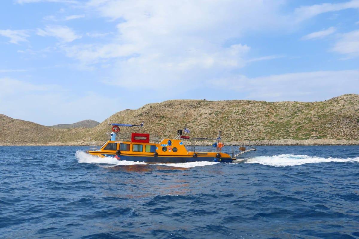 Kos glass bottom boat: an orange and navy glass-bottom speedboat with a red roof box and a small Greek flag throwing white foam as it speeds over deep blue water past a dry brown hilly island coast under a pale blue sky with soft clouds