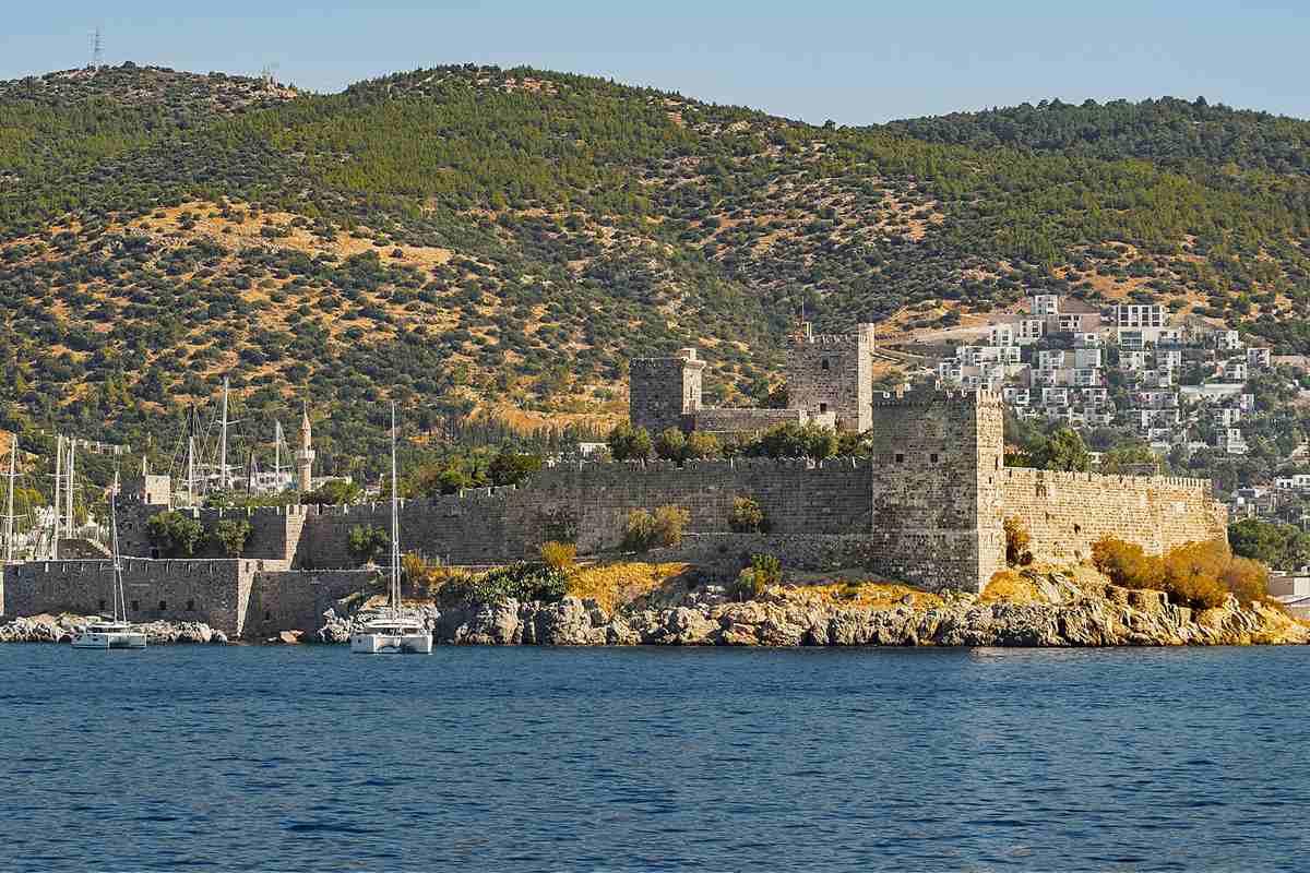 Kos to Bodrum day trip: the grey stone walls and square towers of Bodrum Castle (the Castle of St. Peter) on a rocky headland above deep blue water, with white sailing yachts moored in front, green hills behind and white houses on the right