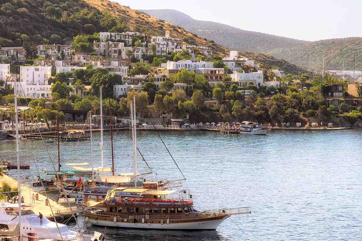 Kos to Bodrum day trip: white houses and hotels climbing a green hillside above the Bodrum shore, with a wooden gulet boat and sailing yachts moored along a quay on calm blue water in warm evening light