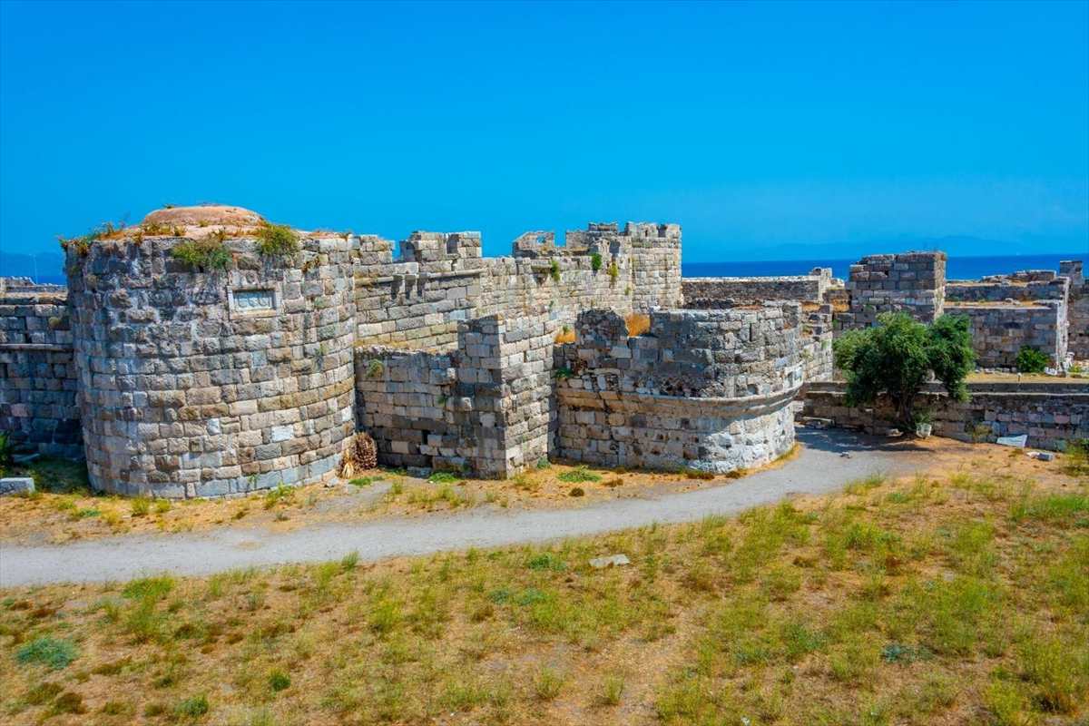 Kos tuk tuk tour: the round stone towers and crenellated walls of Neratzia Castle in Kos Town, with a gravel path curving past dry grass and a green olive tree below a bright blue sky and the sea on the horizon