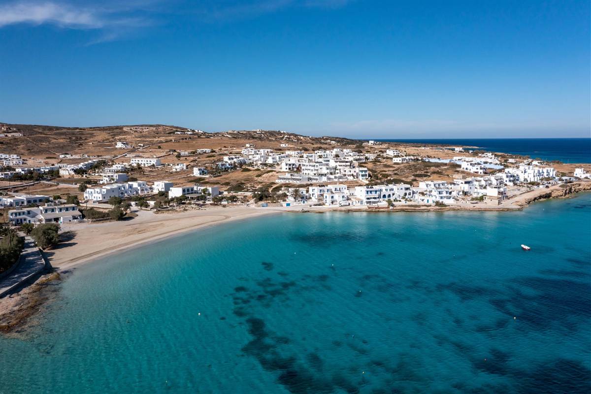 Koufonisia day trip from Naxos: white Cycladic houses of a village on low golden hills behind a curved sandy beach and a wide bay of clear turquoise water under a deep blue sky