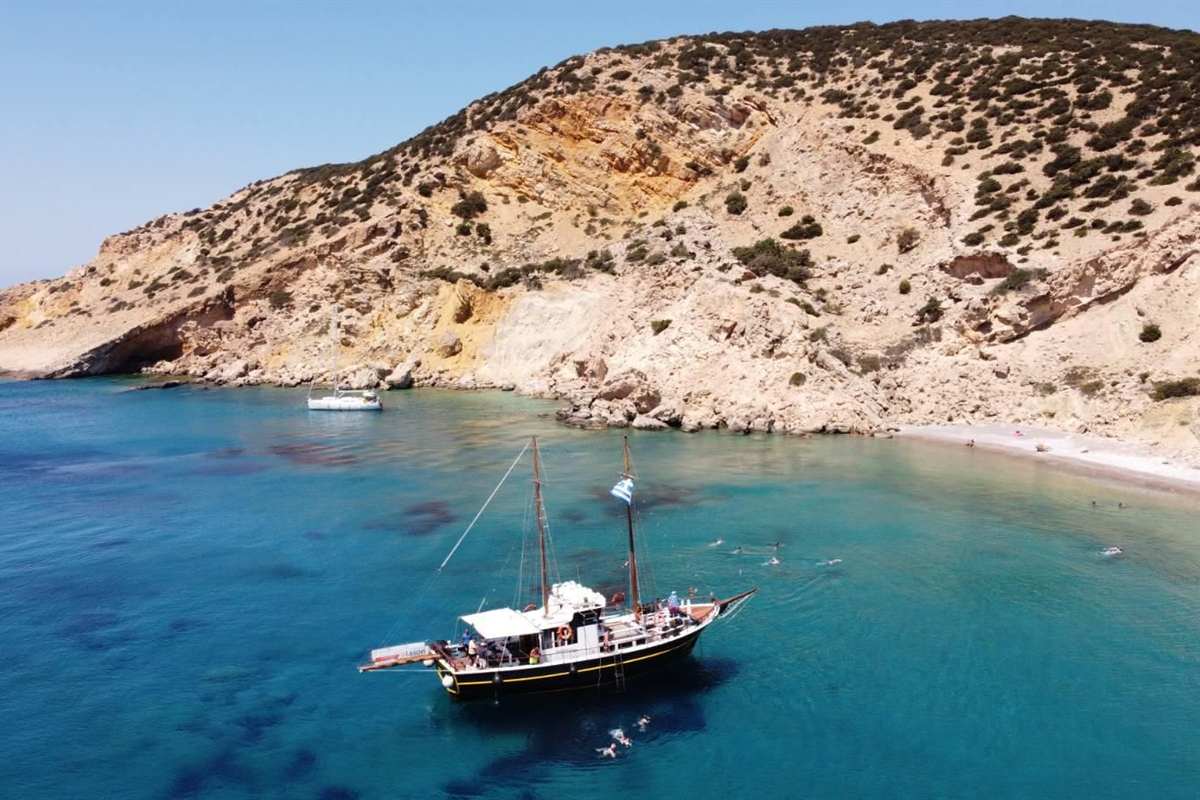 Koufonisia day trip from Naxos: a black-hulled traditional wooden cruise boat with two masts, a white deckhouse and a Greek flag at anchor in a calm blue-green cove below dry golden hills, with a pale beach on the right