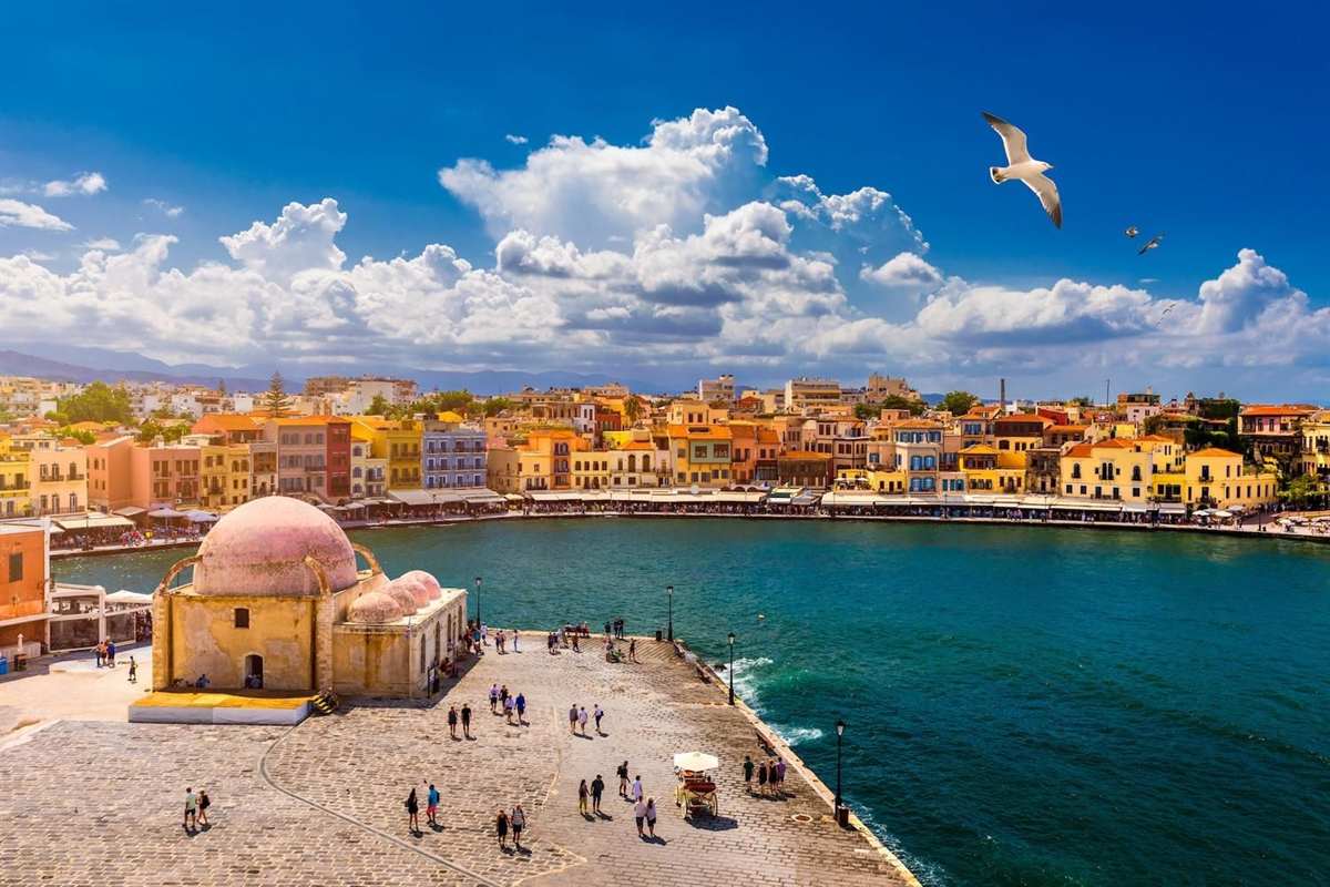 Kournas Lake day trip from Rethymno: the old harbour of Chania from the pier, with the pink domed Kucuk Hasan Pasha mosque on the left, a row of yellow, orange and pink waterfront houses across the turquoise water, a few small groups of visitors walking on the paved quay and a seagull flying under big white clouds