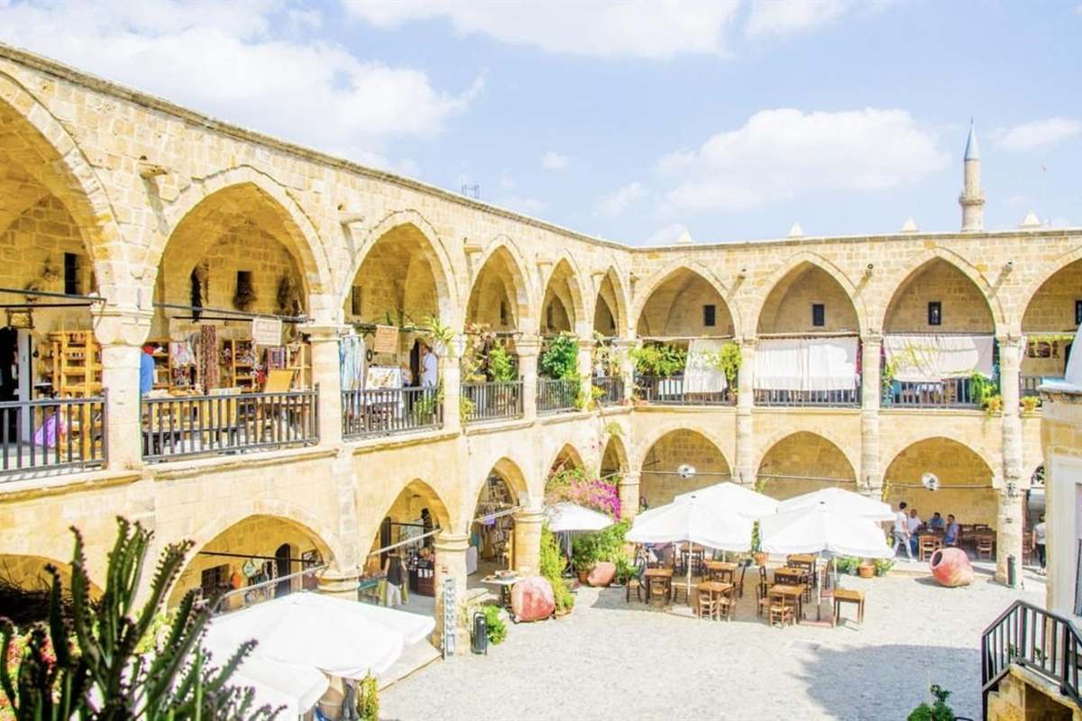 Kyrenia and Nicosia day trip from Ayia Napa: the two-storey courtyard of Buyuk Han in North Nicosia, with pale yellow stone pointed arches, wooden balconies, small craft shops, white parasols over cafe tables and a slim minaret behind, under a blue sky