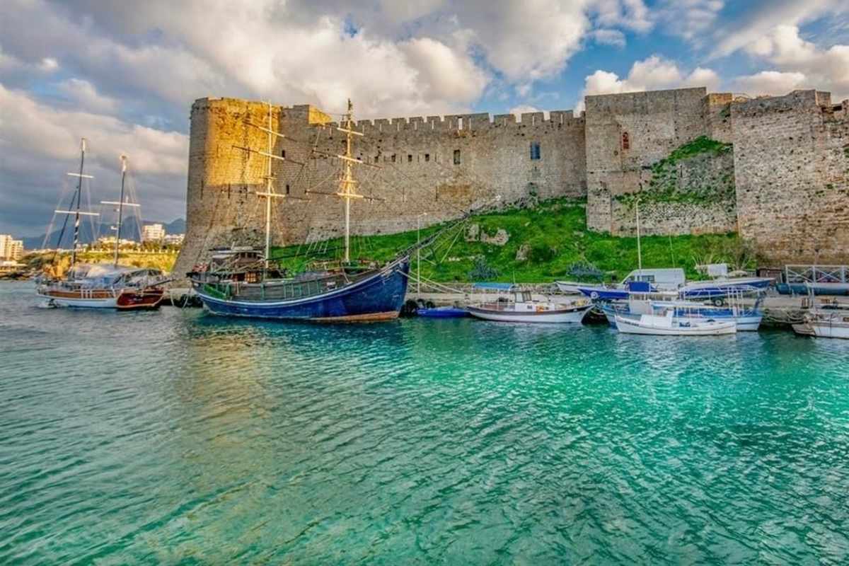 Kyrenia and Nicosia day trip from Ayia Napa: the grey stone walls and towers of Kyrenia Castle above turquoise harbour water, with a dark blue wooden ship with tall masts, a two-masted sailboat and small white fishing boats moored in front, under a bright sky with large clouds