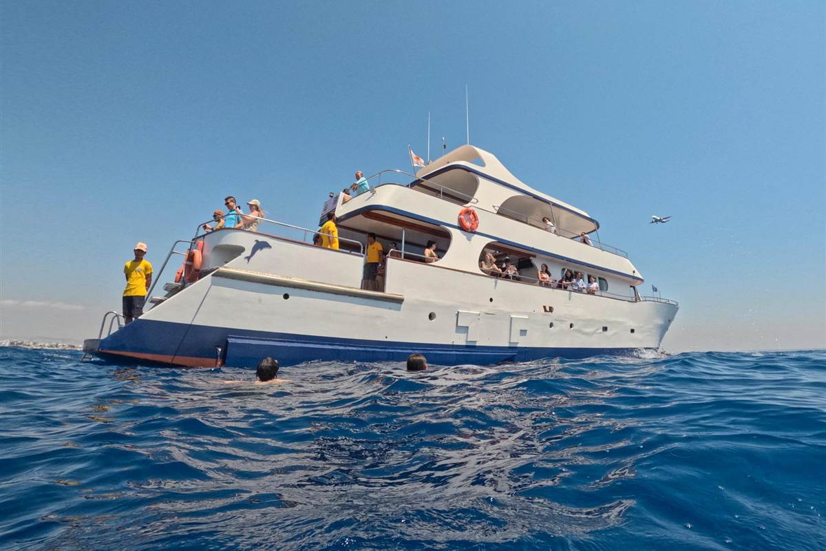 Larnaca snorkeling cruise: a white and navy cruise boat with passengers and yellow-shirted crew on its decks floats at a swim stop on deep blue water while a passenger plane flies low in a clear blue sky and swimmers bob beside the hull