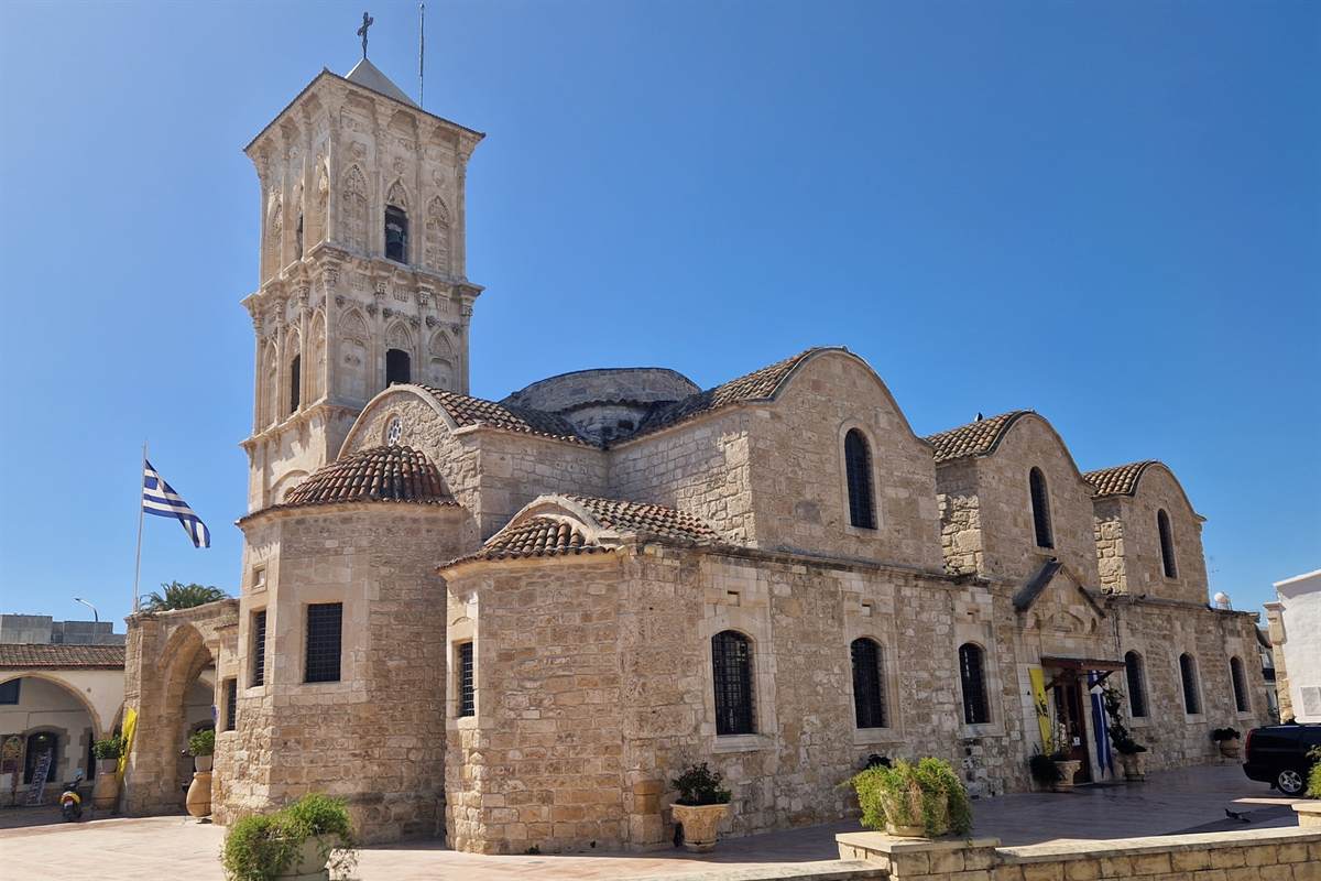 Larnaca sunset cruise: the sandstone bell tower and curved tiled roofs of the Church of Saint Lazarus in Larnaca stand in full view under a clear deep blue sky, with a Greek flag on a pole at the left and a paved square in front