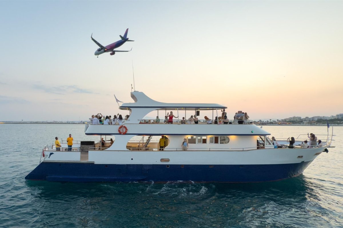 Larnaca sunset cruise: a white and navy two-deck cruise boat with passengers on its decks sails on calm teal water at dusk while a passenger plane flies low overhead, with the Larnaca shoreline and a peach and pale blue sky behind