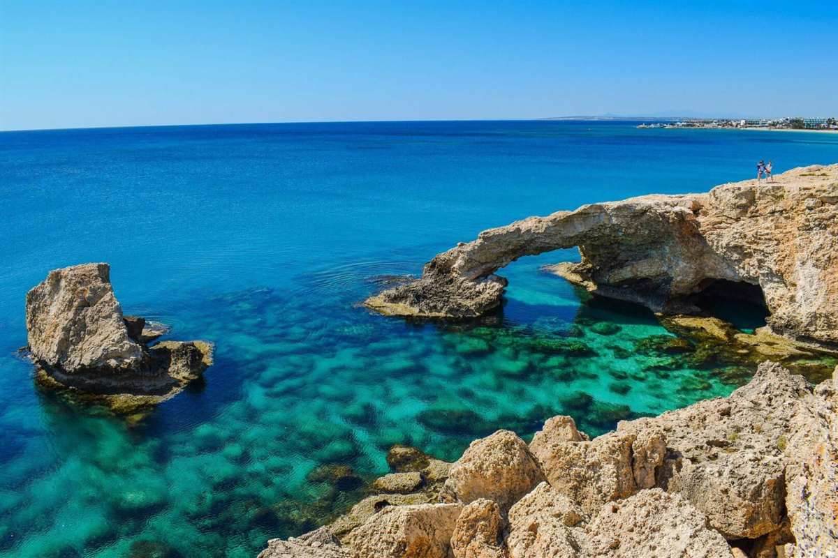Larnaca to Protaras boat safari: a pale limestone sea arch, the Love Bridge near Cape Greco, over clear turquoise water with a rock stack on the left, a rocky shore in the foreground and a blue sky above