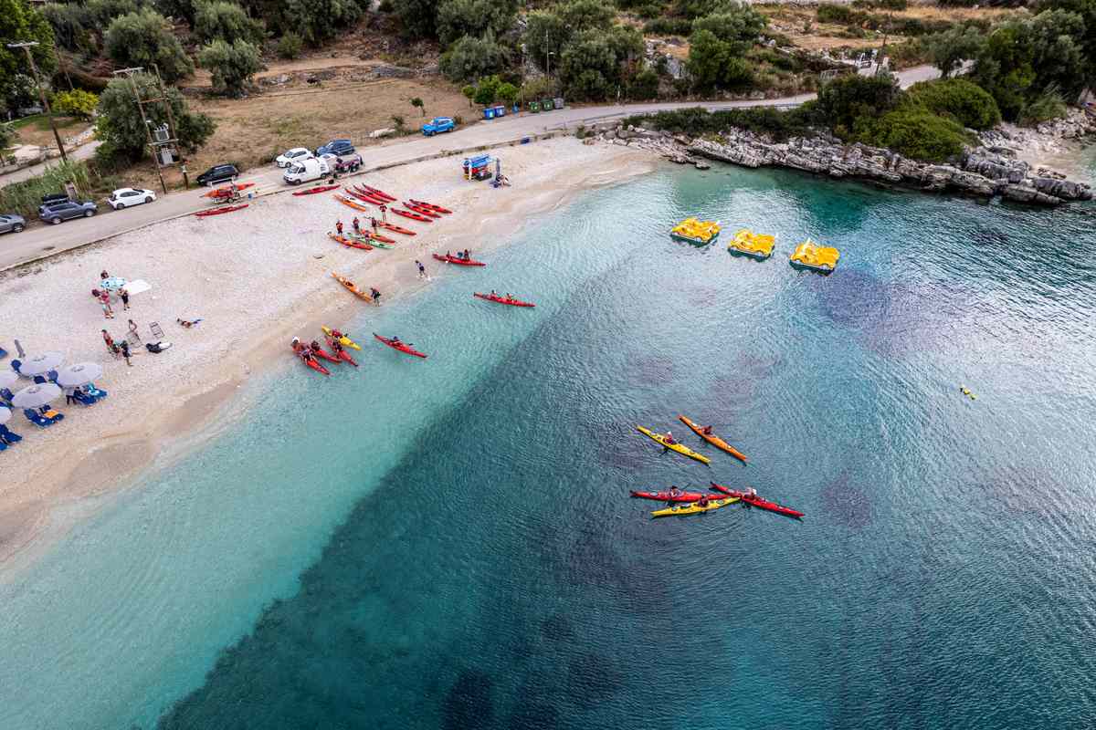Lefkada sea kayak: an aerial view of red and yellow sea kayaks lined up on a white pebble beach and paddling out over turquoise water, with yellow pedal boats, beach umbrellas and olive trees along the coast road
