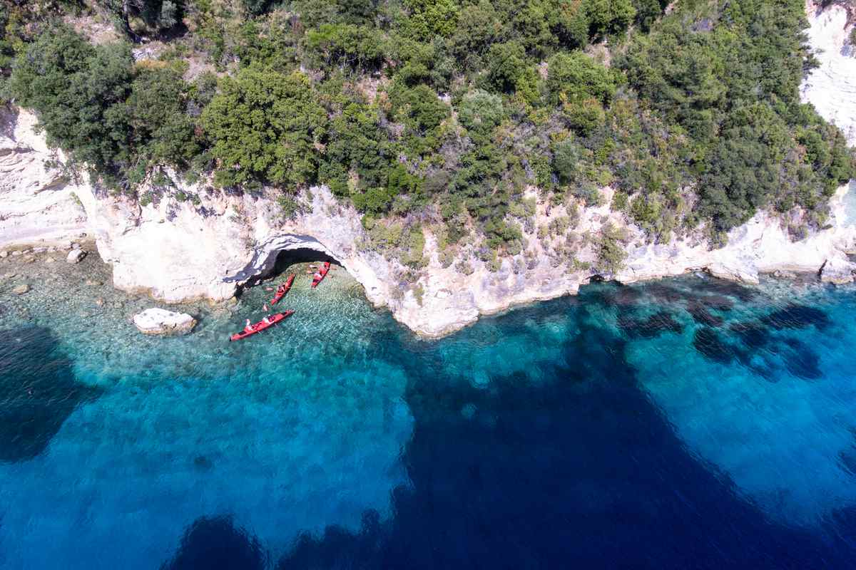 Lefkada sea kayak: an aerial view of red sea kayaks beside a small rock arch cave in pale limestone cliffs topped with green shrubs, above clear turquoise water that turns deep blue offshore