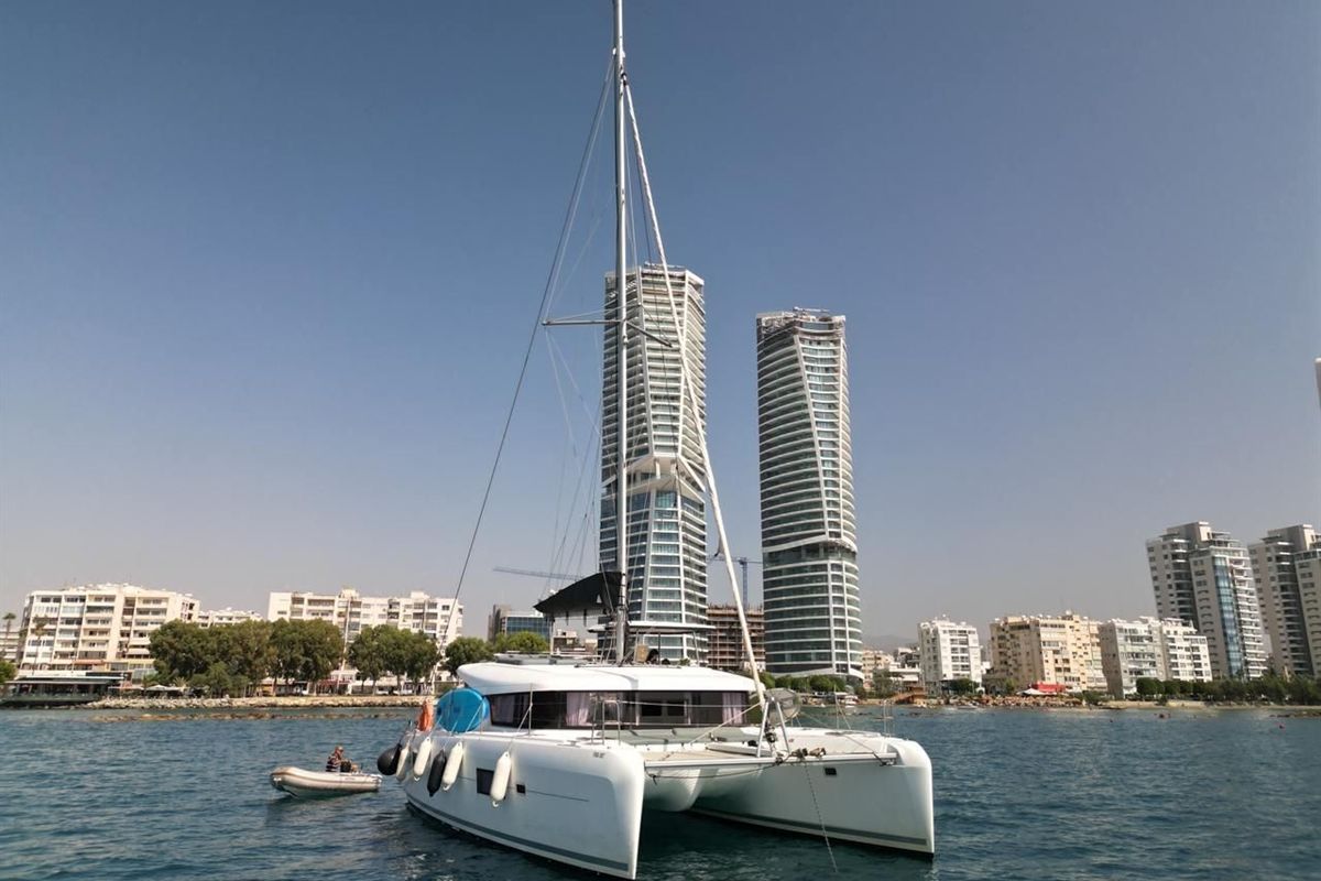Limassol catamaran cruise: a white catamaran with a tall mast and a small grey dinghy beside it floats on calm blue water in front of the Limassol seafront, with two tall glass towers and white apartment blocks behind it under a clear blue sky