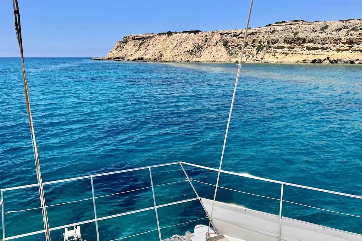 Limassol catamaran cruise: a pale limestone headland with a flat scrubby top rises above bright blue water, seen past the white bow railings and mast ropes of a catamaran under a clear blue sky