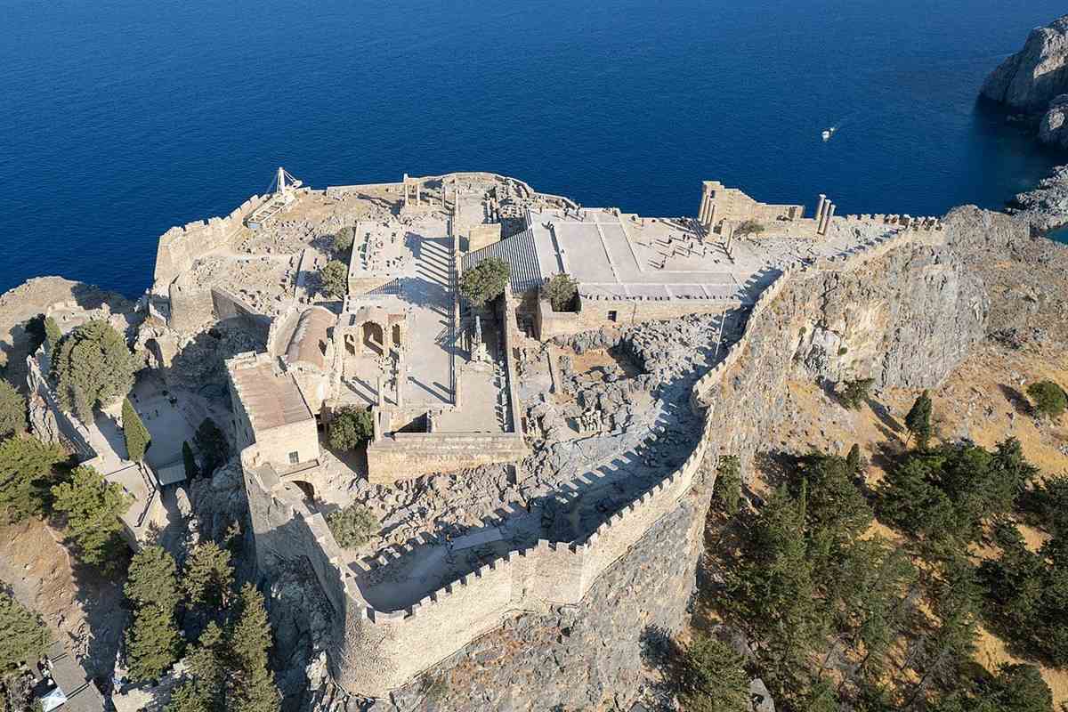 Lindos Acropolis tickets: aerial view of the Acropolis of Lindos, with crenellated stone walls curving around the rocky hilltop, a paved terrace and ruins inside the walls, standing columns at the cliff edge and deep blue sea beyond