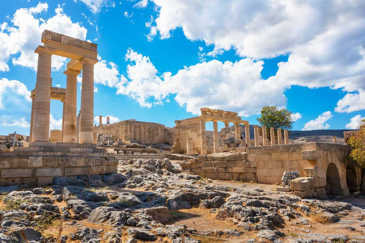 Lindos Acropolis tickets: tall Doric columns carrying a stone beam on a high stone base, a smaller colonnade with an entablature to the right and rocky ground in front, under a bright blue sky with white clouds